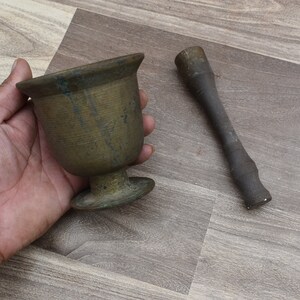 Könnte beinhalten: Ein Mörser und Stößel aus Messing. Der Mörser ist eine schalenförmige Schüssel mit einem abgewinkelten Rand und einem runden Boden. Der Stößel ist ein langer, zylindrischer Stab mit einem abgerundeten Ende.