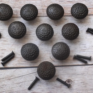 May include: A collection of ten dark gray, textured cabinet knobs with a golf ball-like pattern, along with several black screws. The knobs are arranged on a light brown wooden surface.