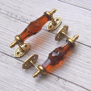 Op de afbeelding: Twee decoratieve kastgrepen met amberkleurige, facetgeslepen glazen handgrepen en sierlijke messingkleurige montageplaten. De hardware is ontworpen voor meubels en voegt een vintage esthetiek toe.