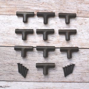 May include: Nine dark bronze T-shaped cabinet pulls and three sets of screws are arranged on a light wood surface. The hardware is likely for drawers or cabinets, offering a rustic or industrial aesthetic.