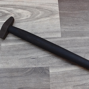 Könnte beinhalten: Ein schwarzer Hammer aus Metall mit einem Holzstiel. Der Hammer liegt auf einer hellgrauen Holzoberfläche.