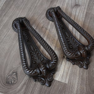 Pode incluir: Dois batentes de porta de ferro fundido preto adornados com desenhos intrincados. Os batentes têm a forma de uma lágrima com uma alça de anel.