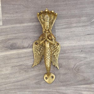 Könnte beinhalten: Ein goldfarbener Türgriff in Form einer Meerjungfrau mit Flügeln und einem kobralike-Kopfschmuck. Die Meerjungfrau hält ihre Hände in einer Gebetsposition zusammen.