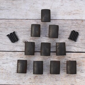 Puede incluir: Un conjunto de diez tiradores de armario cilíndricos de color gris oscuro y dos juegos de tornillos negros dispuestos sobre una superficie de madera clara. Los tiradores tienen un aspecto texturizado e industrial.