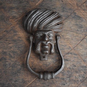 Könnte beinhalten: Ein schwarzer Türklopfer aus Gusseisen in Form eines Männerkopfes mit Turban. Der Türklopfer hat ein detailliertes Gesicht mit einem Schnurrbart und Bart.