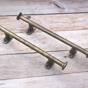 May include: Two antique brass door pulls with a cylindrical bar and rounded ends. Each pull is mounted on a small, rectangular base with two screw holes. The hardware is set against a light-colored, wooden background.