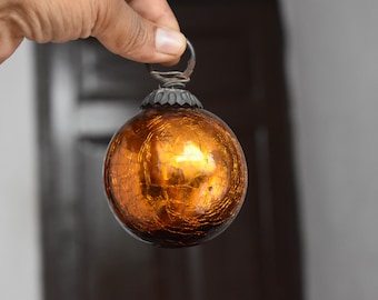 Puede incluir: Un pequeño adorno de vidrio redondo y naranja con un acabado metálico y una textura craquelada. El adorno tiene una tapa de metal plateado con un lazo para colgar.