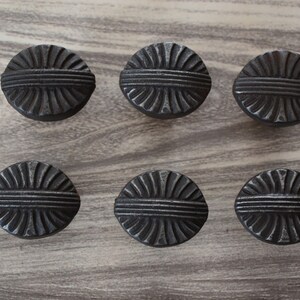 Puede incluir: Seis pomos de armario redondos de color gris oscuro con un diseño texturizado en forma de sol. Los pomos están dispuestos en dos filas de tres sobre una superficie clara con vetas de madera. Estos pomos decorativos son adecuados para muebles o decoración del hogar.