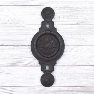 Op de afbeelding: Een zwarte deurklopper van gietijzer met een bloemmotief. De klopper heeft een ronde handgreep en decoratieve elementen aan de boven- en onderkant. De deurklopper is gemonteerd op een wit houten oppervlak.