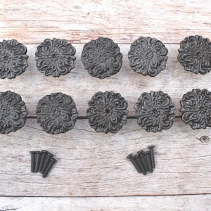Puede incluir: Un juego de diez pomos de armario de color gris oscuro con forma de flor y tornillos a juego. Los pomos tienen un diseño detallado en relieve y están dispuestos sobre una superficie de madera clara. Ideal para añadir un toque decorativo a los muebles.