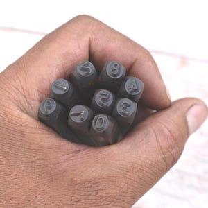 Könnte beinhalten: Nahaufnahme einer Hand, die einen Satz Metall-Zahlenstempel hält. Die Stempel sind dunkelgrau und haben die Zahlen von 0 bis 9. Der Hintergrund ist eine hellfarbige Holzoberfläche.