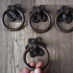 以下が含まれることがあります： 装飾的な花のデザインが施された、アンティーク調の黒い金属製のリングプルが4つ。リングプルは木製の表面にあります。