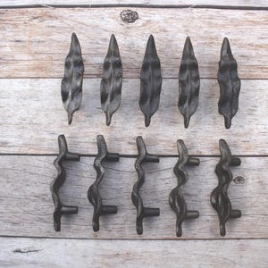Puede incluir: Una colección de diez piezas de herrajes de metal oscuro para armarios. La fila superior presenta seis tiradores en forma de hoja, mientras que la fila inferior tiene cuatro tiradores ondulados. Los herrajes están dispuestos sobre una superficie de madera clara.