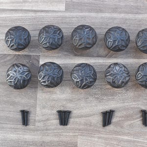 Peut inclure: Douze boutons de porte en métal noir avec un motif de nœud celtique. Les boutons sont disposés en deux rangées de six, chaque rangée étant décalée par rapport à l'autre. Les boutons sont sur une surface en bois brun clair.
