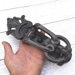 May include: Ornate, dark gray metal drawer pull with a decorative floral and scroll design. The handle is a large, curved loop. The hardware is held in a hand against a white wood background.