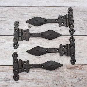 May include: Four black metal T-hinges with a textured, hammered finish. Each hinge features a pointed, spear-shaped end and a rectangular mounting plate with multiple pre-drilled screw holes, designed for heavy-duty door or gate hardware applications.