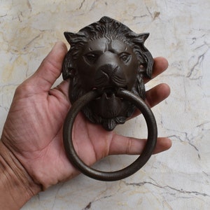 Puede incluir: Un llamador de puerta de cabeza de león de hierro fundido con un asa circular. El llamador es marrón y tiene un diseño detallado.