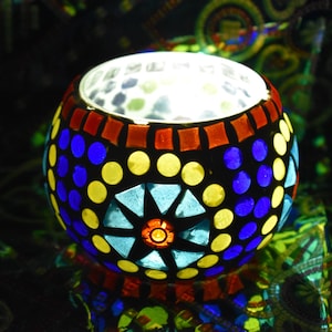 May include: A round glass candle holder with a mosaic design. The holder is made of small, colourful glass tiles in shades of blue, yellow, red, and green. The holder is lit from within, creating a warm glow.