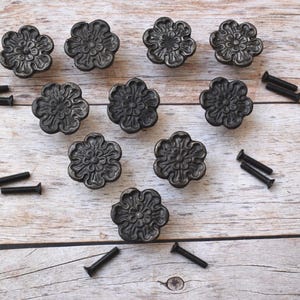 Könnte beinhalten: Neun dunkelgraue, blütenförmige Schrankknöpfe mit passenden Schrauben, angeordnet auf einer hellen Holzoberfläche. Die Knöpfe haben detaillierte Blumenmuster und sind aus Metall. Ideal für Möbelrestaurierung oder DIY-Projekte.