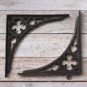 Op de afbeelding: Twee zwarte metalen plankdragers met een L-vormig ontwerp en decoratieve uitsparingen. Elke drager toont een vierpaspatroon in de hoeksteun, wat zorgt voor een structureel frame met een matte afwerking en een opengewerkt uiterlijk.