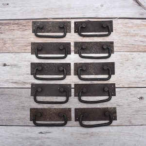 May include: A set of ten rectangular metal drawer pulls with a dark, weathered finish. Each pull features a flat, textured mounting plate with two screw holes and a hinged, U-shaped handle attached to two circular pivot points on the plate.