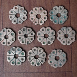 Puede incluir: Doce abalorios de metal de color plateado en forma de flor con un agujero en el centro. Cada abalorio tiene un diseño circular con un centro elevado y un anillo de círculos más pequeños alrededor del borde.