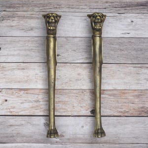 May include: A pair of long, vertical door handles crafted from antique brass-toned metal. Each handle features a detailed, snarling panther head at the top and a stylized paw design at the base, with a smooth, slender shaft connecting the two ends.