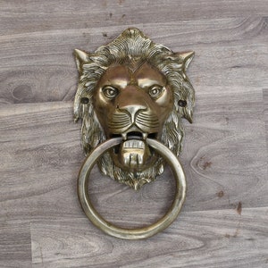 May include: A brass lion's head door knocker with a circular ring. The knocker is mounted on a wooden surface.