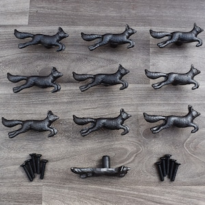 Könnte beinhalten: Zehn schwarze Metall-Schubladenknöpfe in Form eines laufenden Fuchses. Die Knöpfe sind in drei Reihen auf einer Holzoberfläche angeordnet.