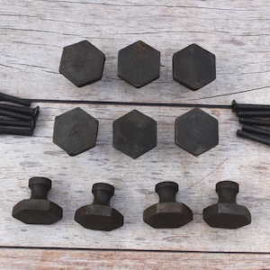 Puede incluir: Un juego de diez pomos hexagonales para armarios en color negro mate oscuro con tapas planas y bases cónicas, acompañados de diez tornillos de montaje de metal negro a juego. Los pomos están dispuestos en filas, mostrando caras y perfiles.