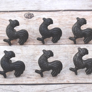 May include: Six black cast iron rooster-shaped cabinet pulls. The pulls have a textured finish and are arranged on a light wood surface. These decorative pulls are ideal for adding a rustic touch to furniture.