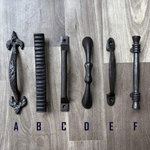 Könnte beinhalten: Sechs schwarze Türgriffe aus Gusseisen in verschiedenen Designs. Zu den Stilen gehören eine Fleur-de-Lis, ein gerippter Stab, ein einfacher Stab, ein geschwungenes Design, ein flacher Griff und ein Griff mit seilartiger Textur. Jeder Griff ist mit einem Buchstaben gekennzeichnet.