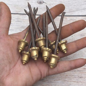 May include: A collection of metal upholstery nails featuring dark, slender, pointed shafts and polished brass-toned, acorn-shaped decorative heads. These hardware fasteners are designed with a distinct conical base and a sharp, straight metal spike.