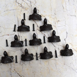 May include: Twelve black cast iron turtle drawer pulls with screws. The pulls are arranged in a triangular pattern on a white marble surface.