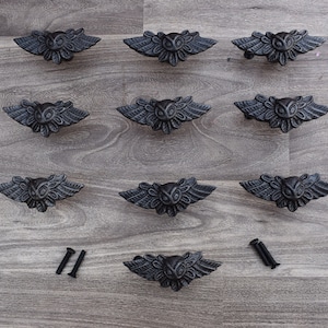 May include: A collection of eleven cast iron owl-shaped cabinet pulls in a dark gray finish. Each pull features an owl with outstretched wings. Two screws are included for installation. The pulls are arranged on a gray wood-grain surface.
