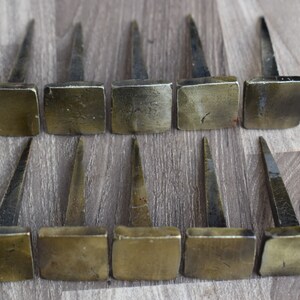 Puede incluir: Doce clavos de cabeza cuadrada con acabado martillado. Los clavos son de metal y tienen un color dorado claro.