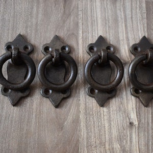 Pode incluir: Quatro puxadores de gaveta de ferro fundido de estilo antigo com uma alça de anel e um design decorativo de flor-de-lis.