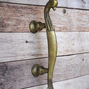 May include: A brass-colored door handle with a peacock design. The handle features a curved body and a bird's head at the top. The handle is mounted on a wooden surface.
