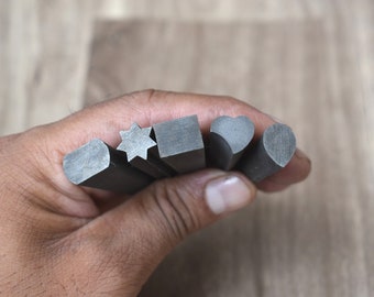 May include: A set of five metal stamps with different shapes, including a heart, a star, a square, and two rounded shapes. The stamps are held in a hand.