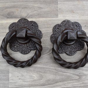 Peut inclure: Deux heurtoirs de porte décoratifs marron foncé. Chaque heurtoir présente une poignée en anneau torsadé et une plaque arrière à motifs floraux. Les heurtoirs sont en métal et sont présentés sur une surface en bois gris clair.
