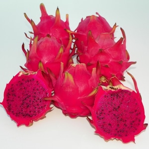 Op de afbeelding: Verschillende felroze drakenvruchten, sommige heel en andere doormidden gesneden om het diepe magenta vruchtvlees met zwarte zaden te onthullen. De vruchten hebben groene en gele stekelige uitsteeksels.