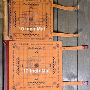 以下が含まれることがあります： 「10 Inch Mat」と「12 Inch Mat」と書かれた2つのタン革製ゲームマット。各マットは、バイキングをテーマにしたイラストとテキストが施されたグリッドデザインが特徴です。マットには固定用のストラップとジッパー付きのエッジが付いています。