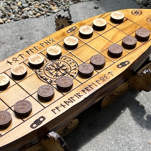 Puede incluir: Juego de mesa de madera con tem&aacute;tica vikinga, con forma de barco. El tablero marr&oacute;n claro presenta una cuadr&iacute;cula, runas talladas y piezas de juego marrones claras y oscuras. El tablero est&aacute; sostenido por patas con forma de drag&oacute;n.