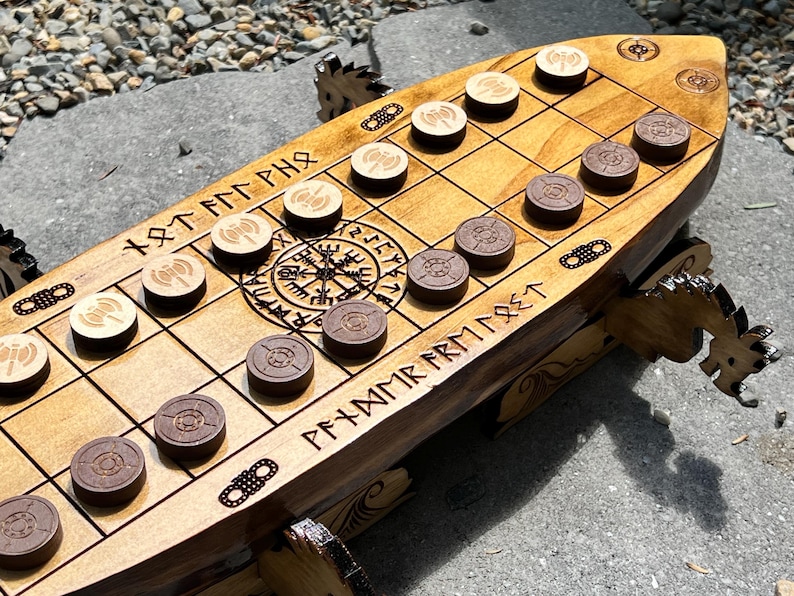 Puede incluir: Juego de mesa de madera con tem&aacute;tica vikinga, con forma de barco. El tablero marr&oacute;n claro presenta una cuadr&iacute;cula, runas talladas y piezas de juego marrones claras y oscuras. El tablero est&aacute; sostenido por patas con forma de drag&oacute;n.