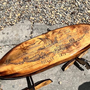 Puede incluir: Una tabla de surf de madera con un dise&ntilde;o grabado. La obra de arte representa un barco vikingo, criaturas marinas y p&aacute;jaros. La tabla est&aacute; soportada por un soporte de madera y presenta el nombre "MUELLER - 2025".