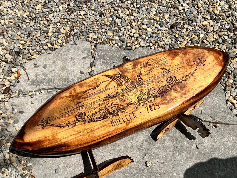 Puede incluir: Una tabla de surf de madera con un dise&ntilde;o grabado. La obra de arte representa un barco vikingo, criaturas marinas y p&aacute;jaros. La tabla est&aacute; soportada por un soporte de madera y presenta el nombre "MUELLER - 2025".