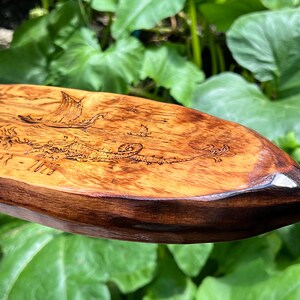 Puede incluir: Pieza decorativa ovalada de madera con una escena n&aacute;utica grabada. La obra de arte representa un barco vikingo, criaturas marinas y el texto "MUELLER - 2025". La madera tiene un tono c&aacute;lido con un borde marr&oacute;n oscuro.