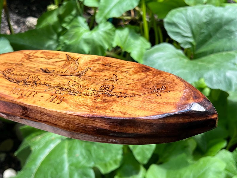 Puede incluir: Pieza decorativa ovalada de madera con una escena n&aacute;utica grabada. La obra de arte representa un barco vikingo, criaturas marinas y el texto "MUELLER - 2025". La madera tiene un tono c&aacute;lido con un borde marr&oacute;n oscuro.