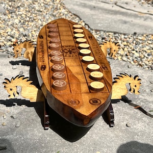 Puede incluir: Un juego de ajedrez vikingo de madera con un tablero en forma de barco y soportes en forma de caballo. El tablero presenta un patr&oacute;n de cuadr&iacute;cula con piezas de juego marrones y beige. El juego est&aacute; sobre una superficie de piedra.