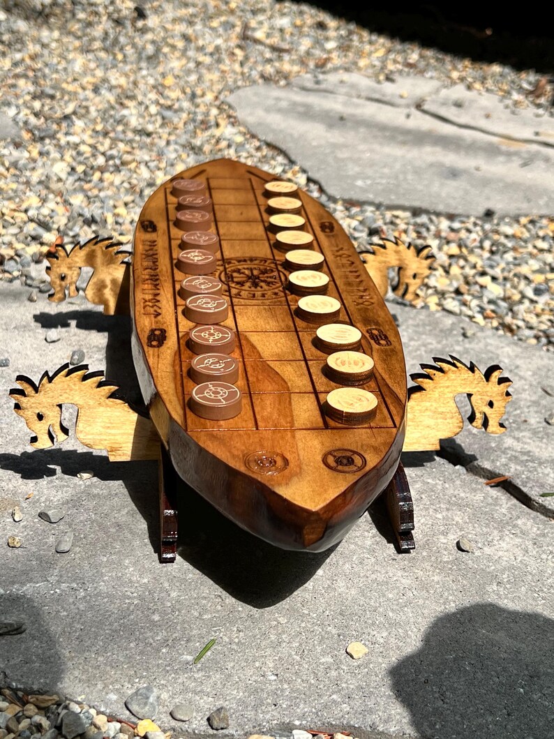 Puede incluir: Un juego de ajedrez vikingo de madera con un tablero en forma de barco y soportes en forma de caballo. El tablero presenta un patr&oacute;n de cuadr&iacute;cula con piezas de juego marrones y beige. El juego est&aacute; sobre una superficie de piedra.